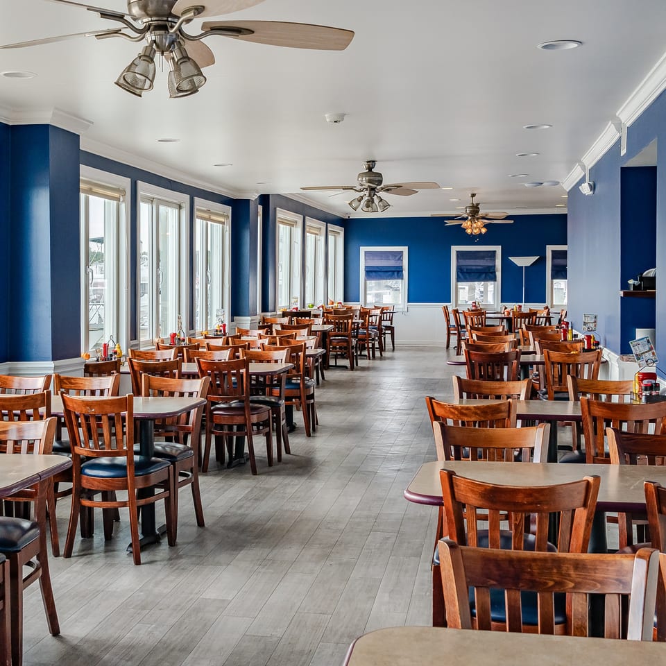 Spacious waterfront restaurant dining room with blue walls, wooden tables and chairs, ceiling fans, and marina views through large windows.