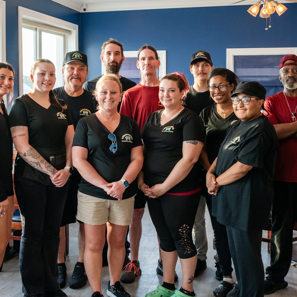 Restaurant staff pose together in two rows inside a bright dining room with blue walls and large windows.