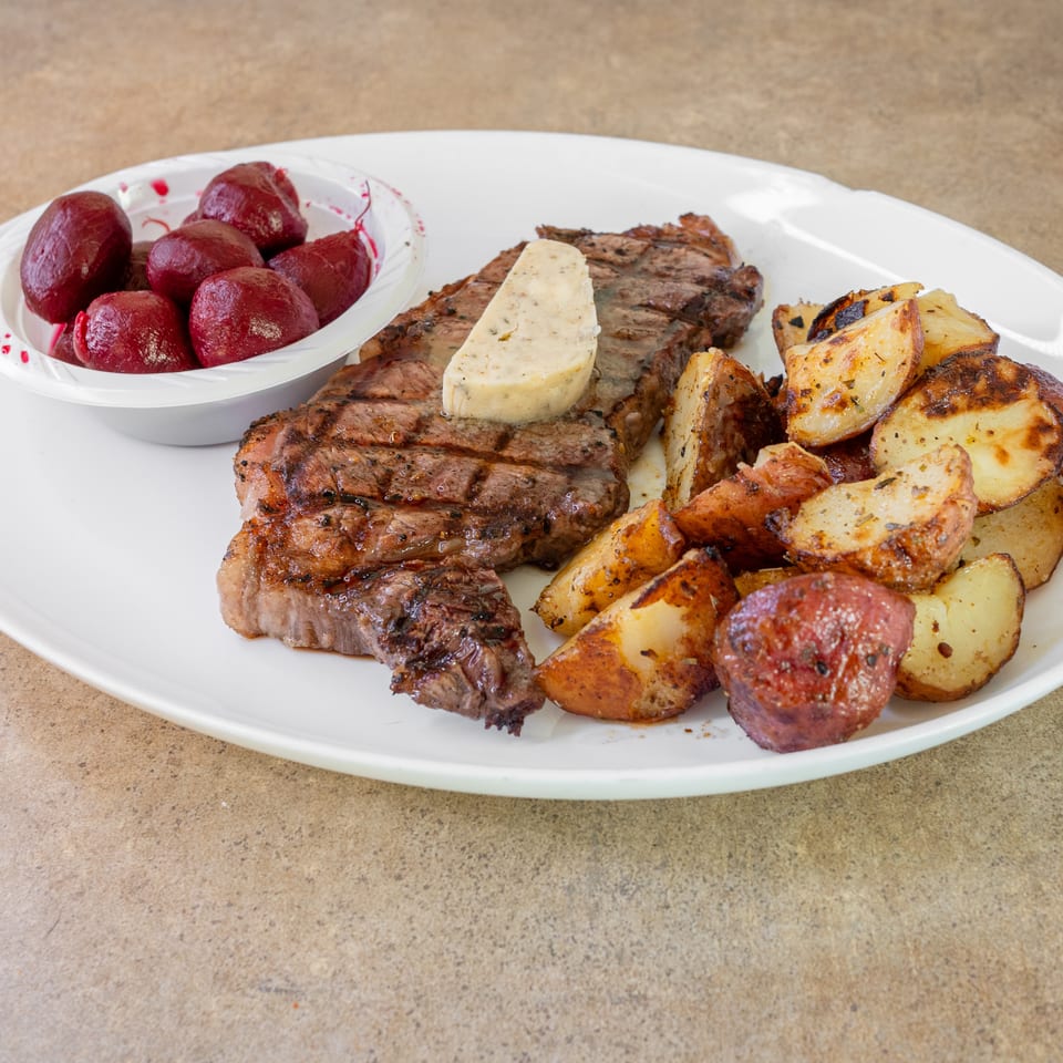 Grilled steak topped with herb butter, roasted potatoes, and whole roasted beets on a white platter.