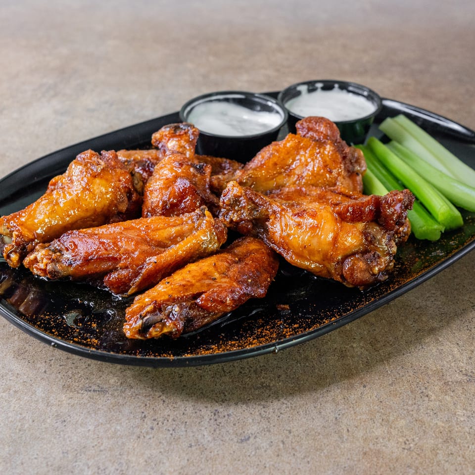 Glazed chicken wings served on a black platter with celery sticks and two cups of creamy dipping sauce.