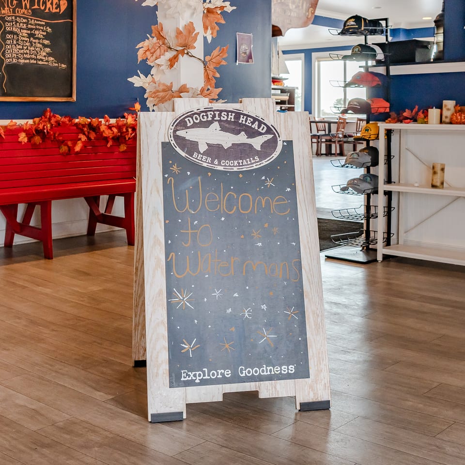 Dogfish Head Beer & Cocktails restaurant interior with a welcome chalkboard, seasonal leaf decorations, red bench, and hat display.