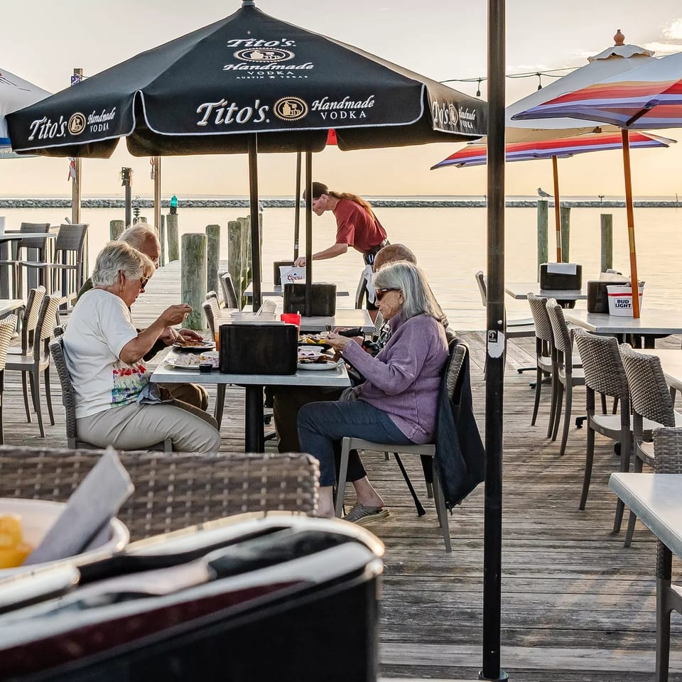 Diners enjoy a waterfront meal on a wooden deck beneath colorful umbrellas overlooking calm water.