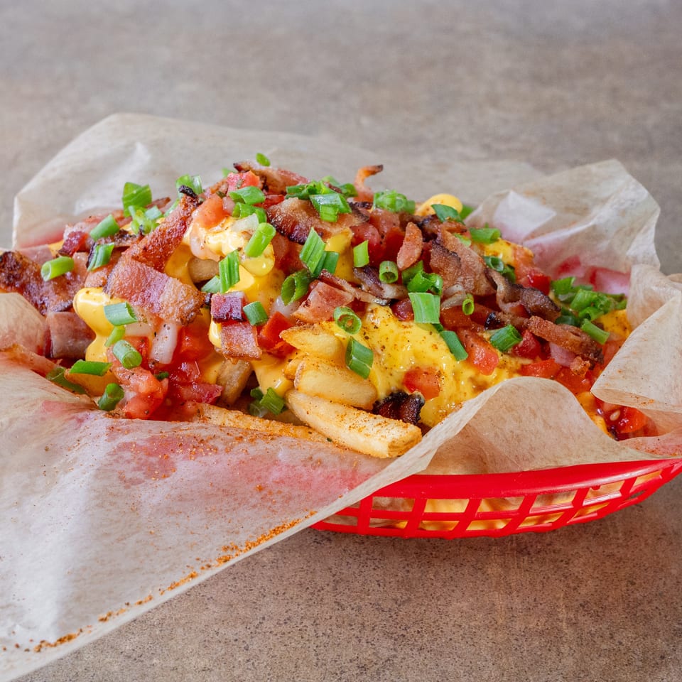 Loaded fries topped with melted cheese, crispy bacon, diced tomatoes, and green onions in a red basket.