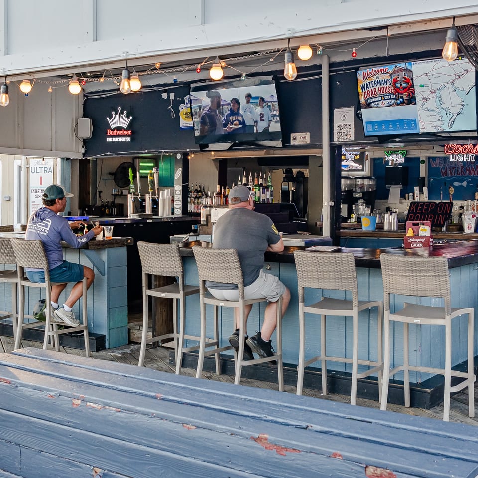 Outdoor waterfront bar with string lights, beer taps, televisions, nautical decor, and two patrons seated at the counter.