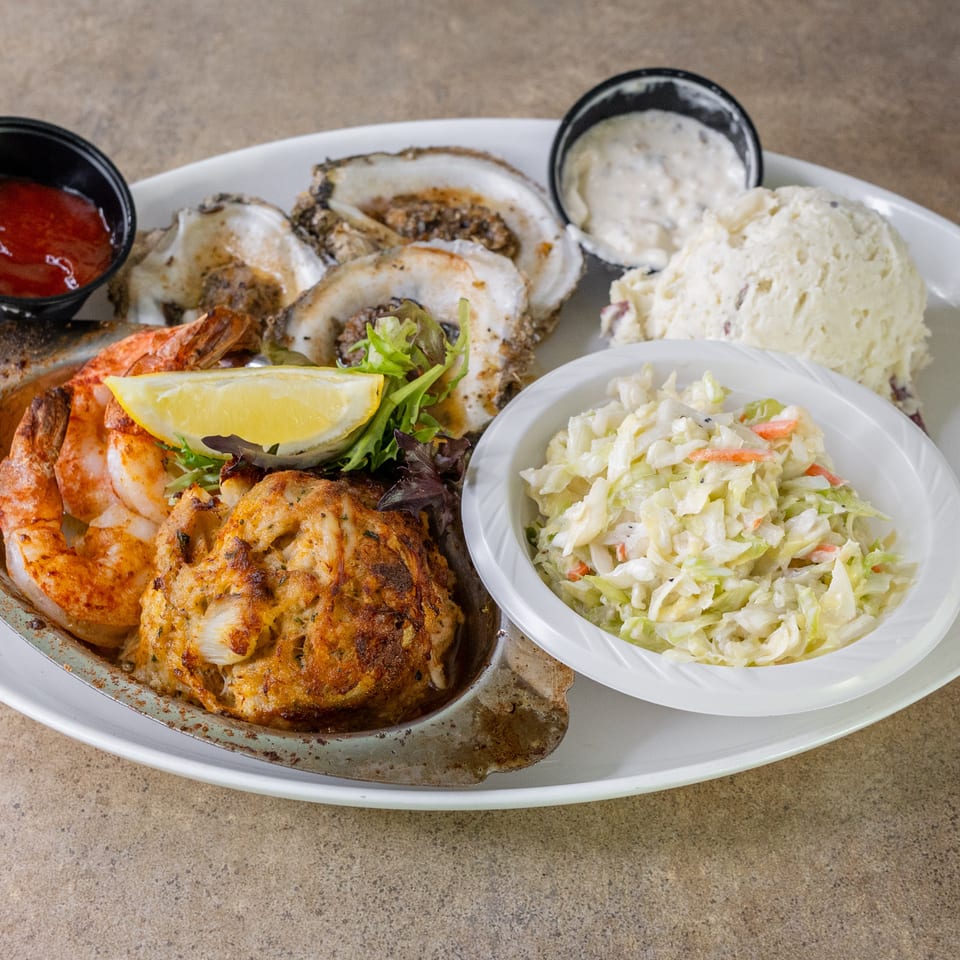 Seafood platter with grilled shrimp, oysters, crab cakes, coleslaw, potato salad, lemon wedge, and dipping sauces.