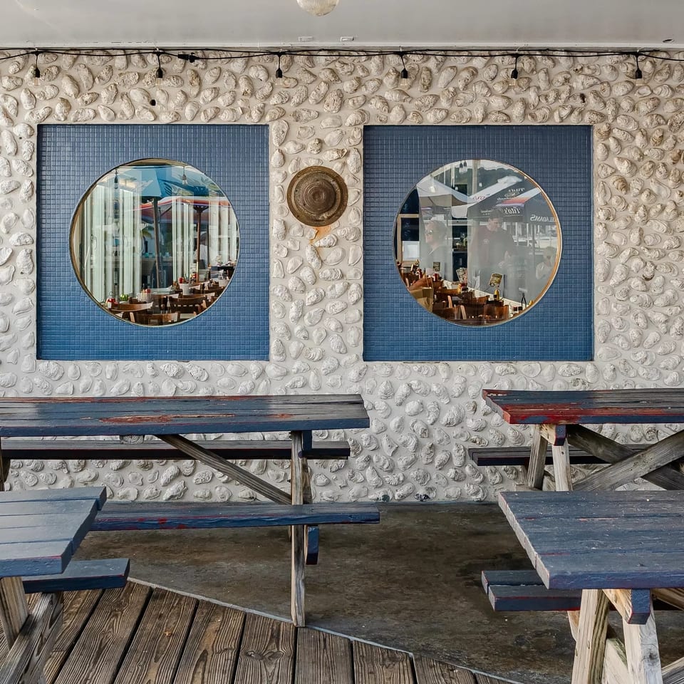 Outdoor restaurant seating with weathered picnic tables, shell-covered walls, and round windows revealing the dining area.